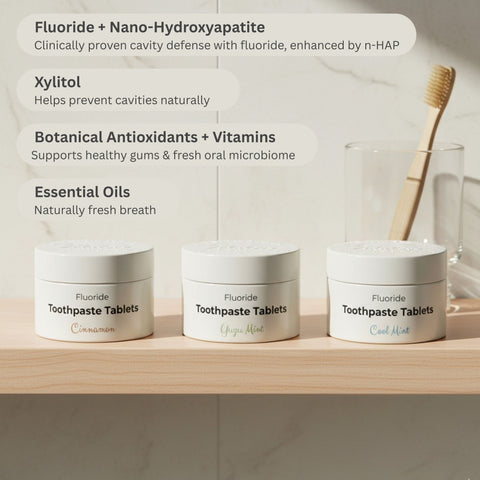 Three containers of fluoride toothpaste tablets on a wooden shelf with a glass of water and toothbrush in the background.