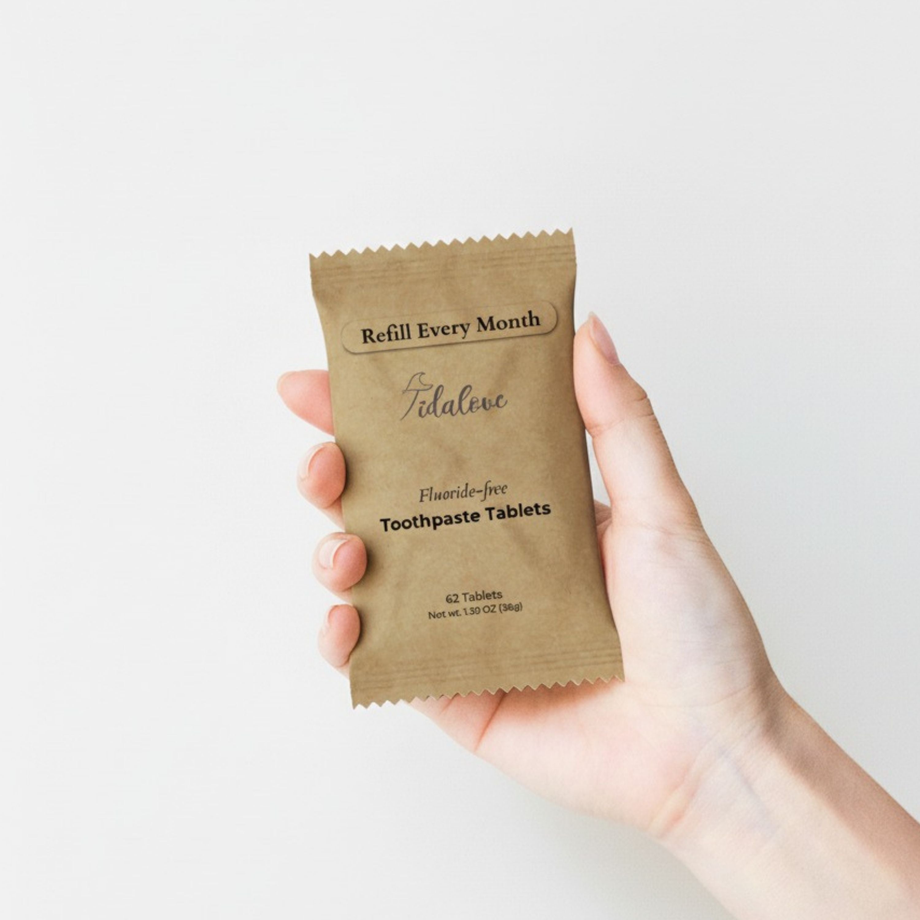 Hand holding a brown package of fluoride-free toothpaste tablets with 'Refill Every Month' text on a light background.