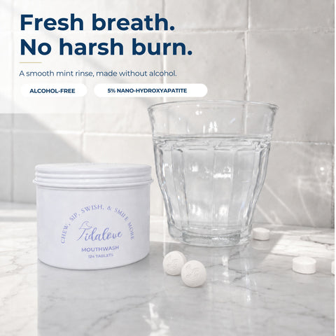 A white tub of Tidalove mouthwash with three tablets beside a clear glass on a marble surface against a tiled wall. Text at the top reads: 'Fresh breath.